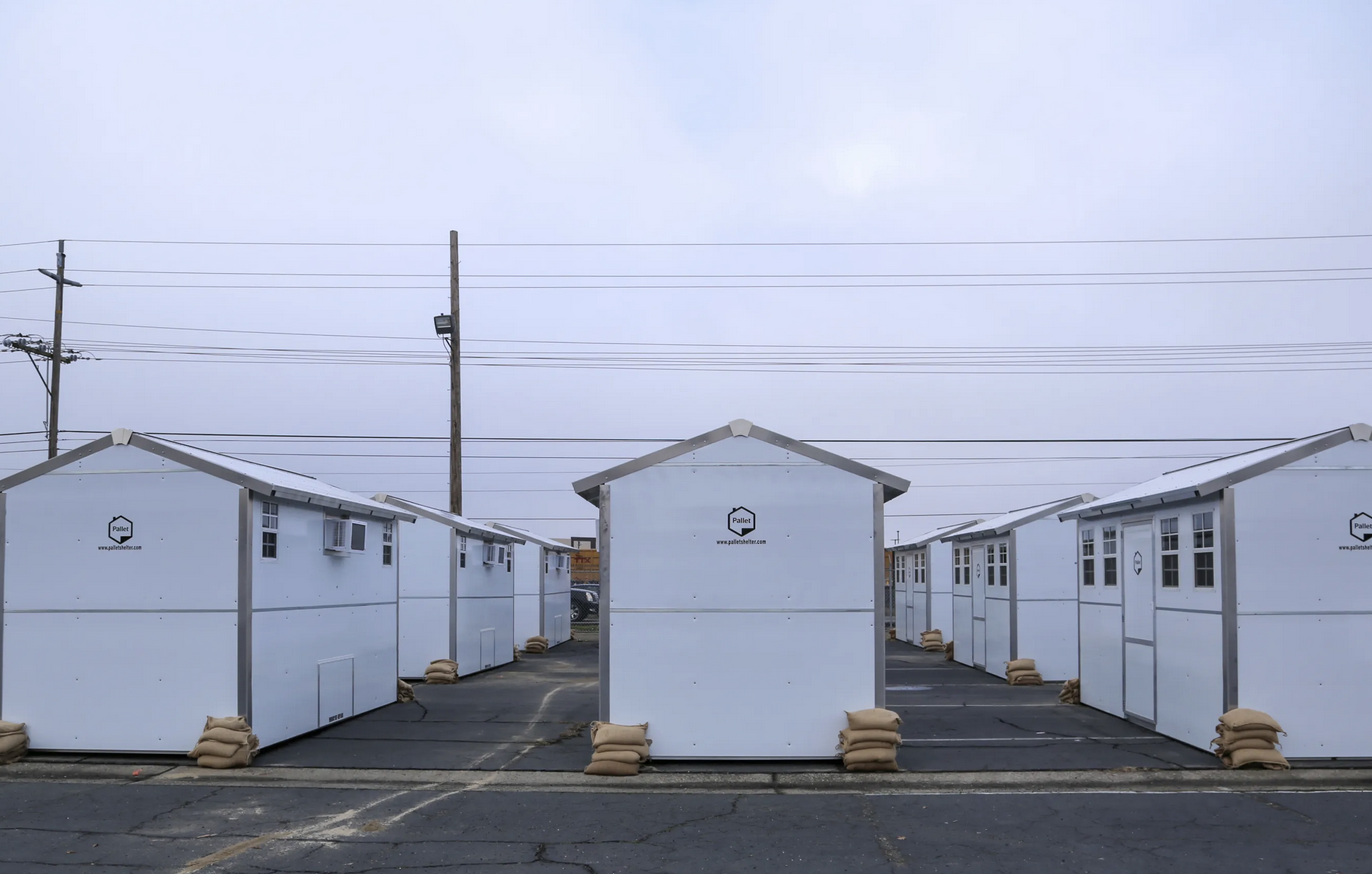 Small white shelters with sandbags in rows on a paved area, under a cloudy sky.
