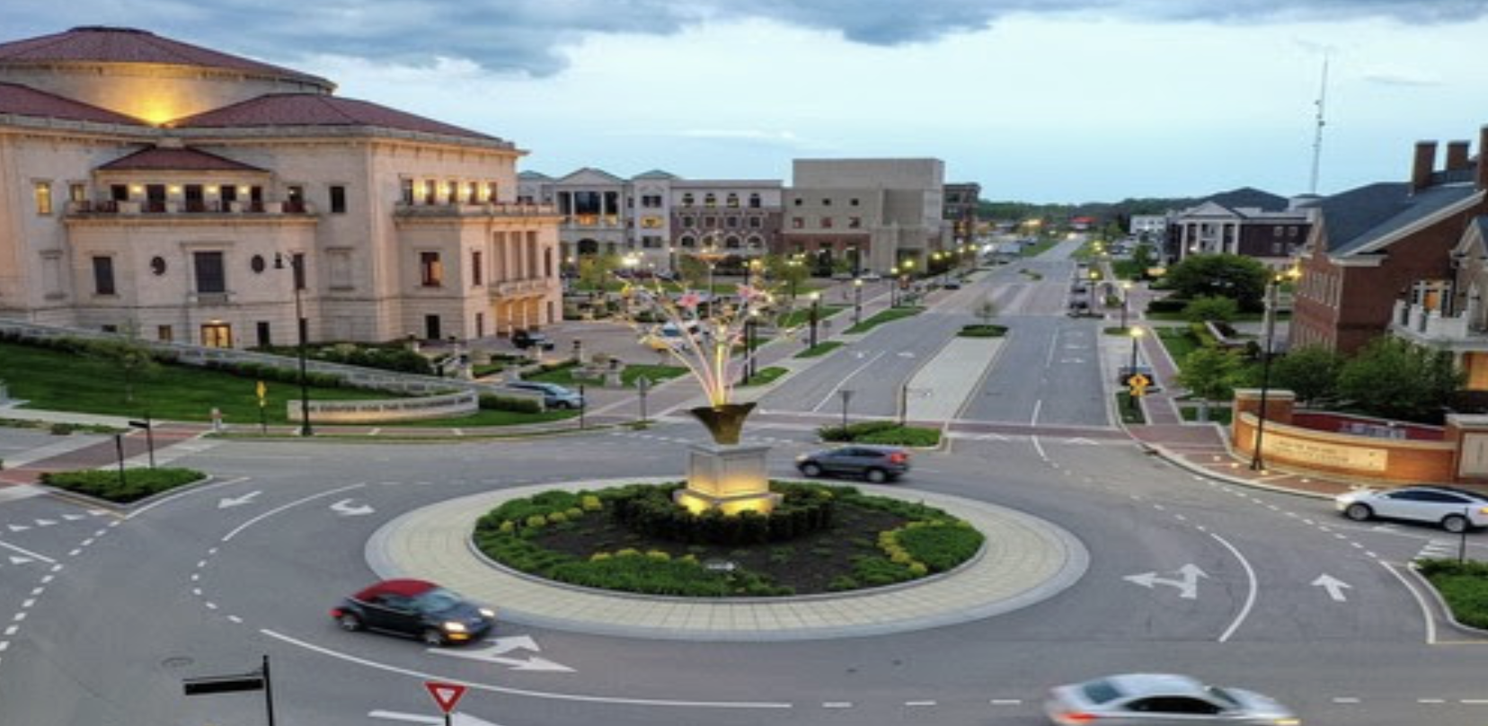 A roundabout with a sculpture at the center, surrounded by buildings and illuminated streetlights.