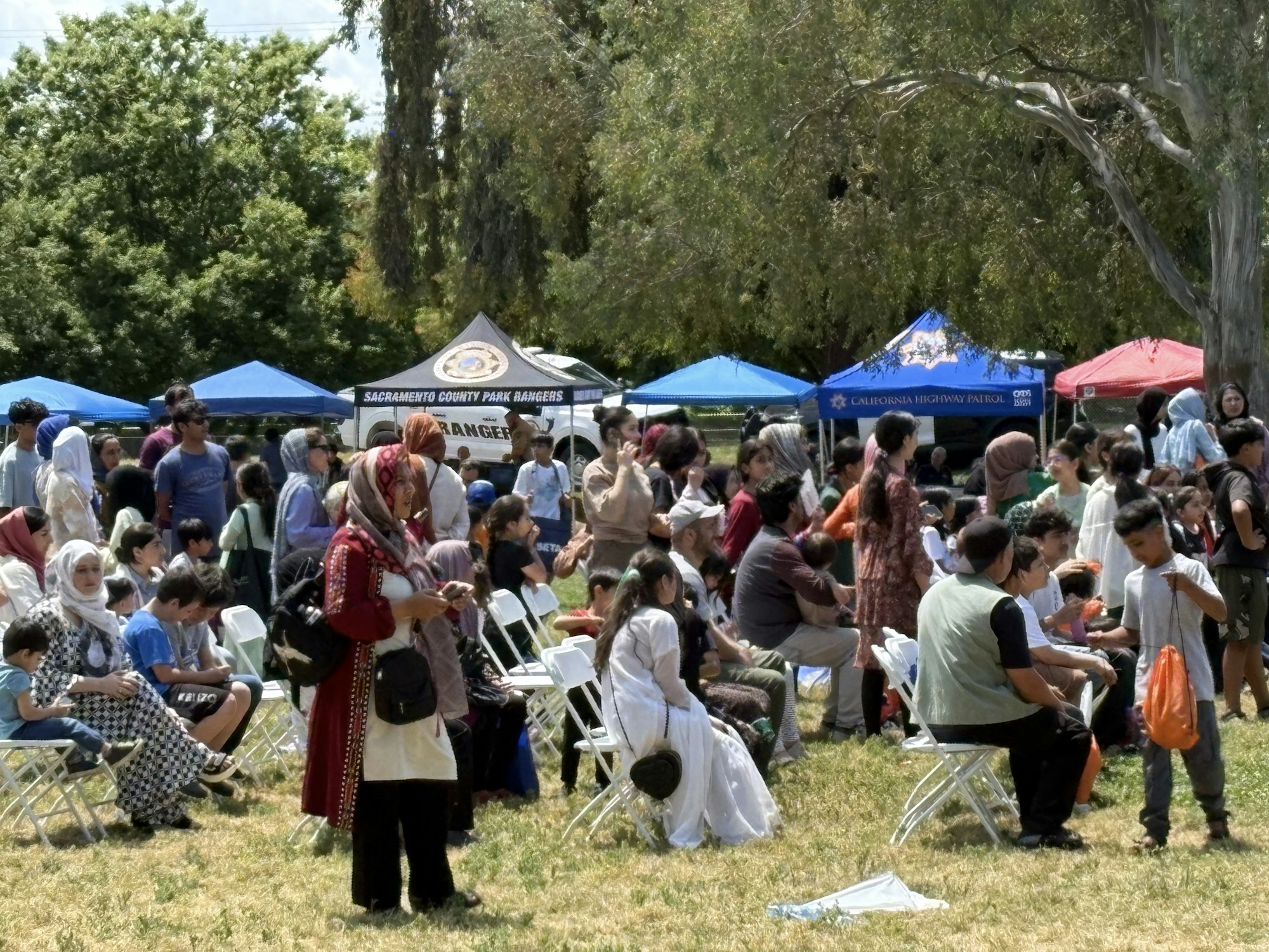 Outdoor gathering with many people, some seated, under trees and tents, including Sacramento County Park Rangers and California Highway Patrol.