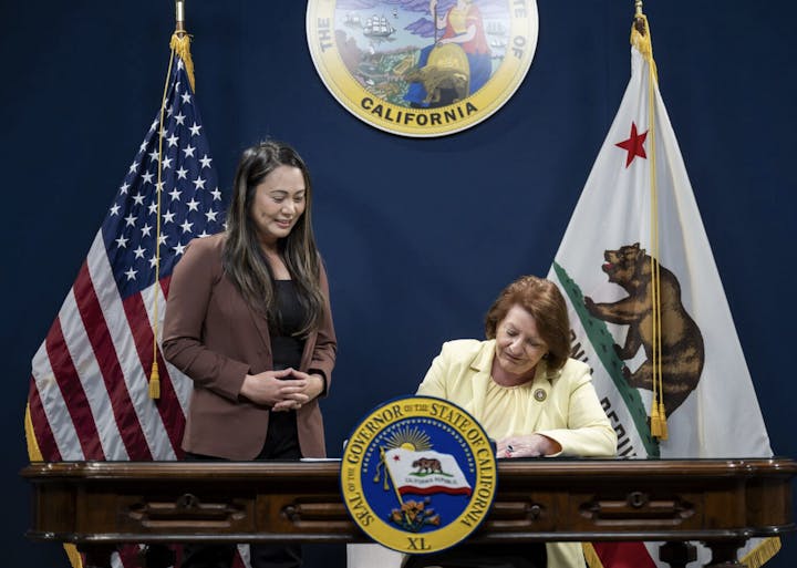 A woman is signing a document at a desk, with flags and a state seal of California in the background.
