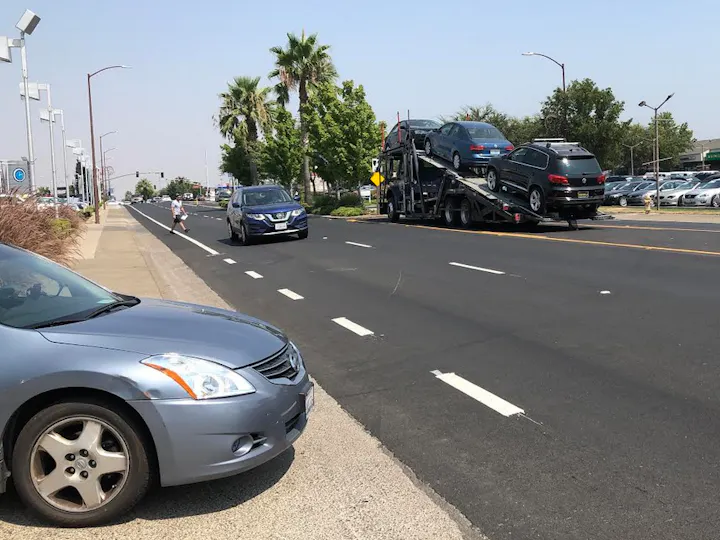 A silver car is parked on the side of a road with a car carrier unloading vehicles nearby, and a person walking on the sidewalk.