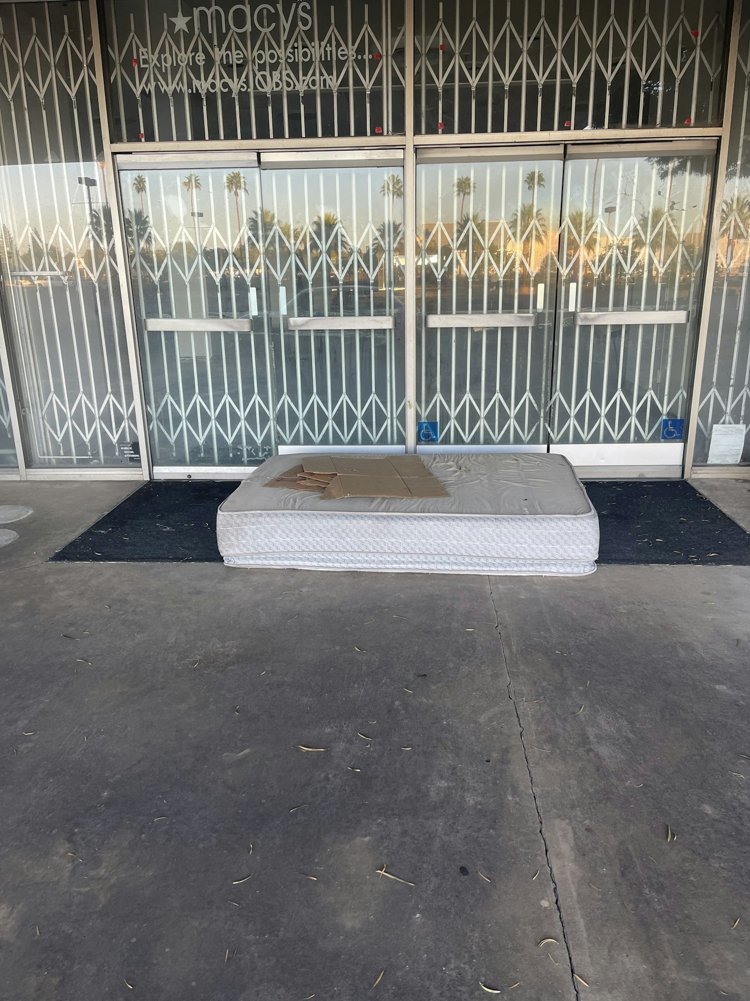 A mattress and cardboard placed on the ground in front of a closed, gated store entrance.