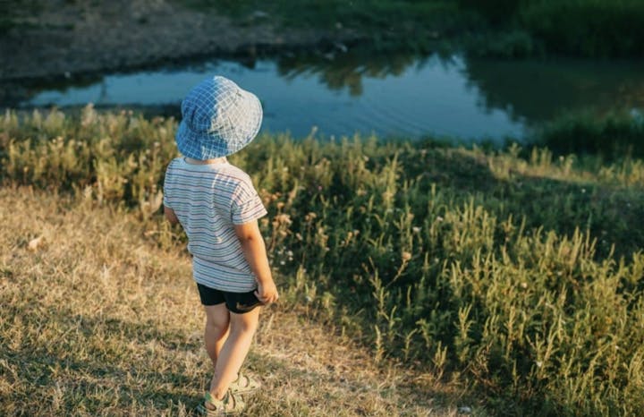 A child wearing a blue hat stands on grass, looking at a small body of water.