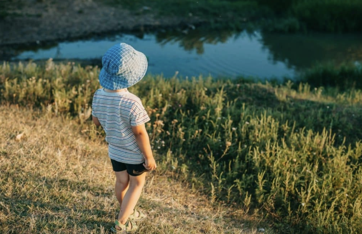 A child wearing a blue hat stands on grass, looking at a small body of water.