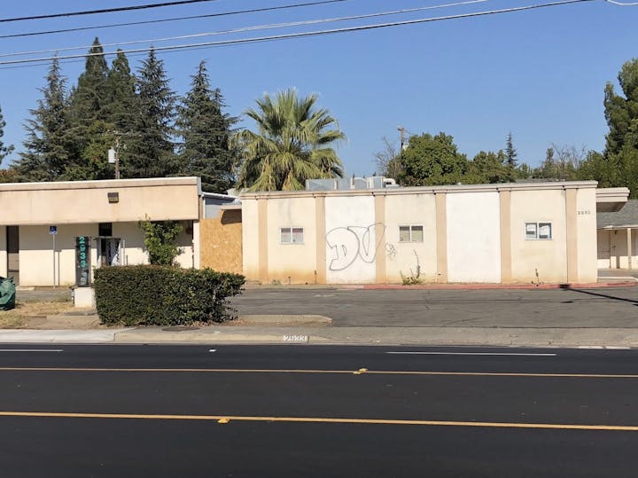 An empty lot with a partially abandoned building, graffiti on a wall, and some trees in the background under a clear blue sky.