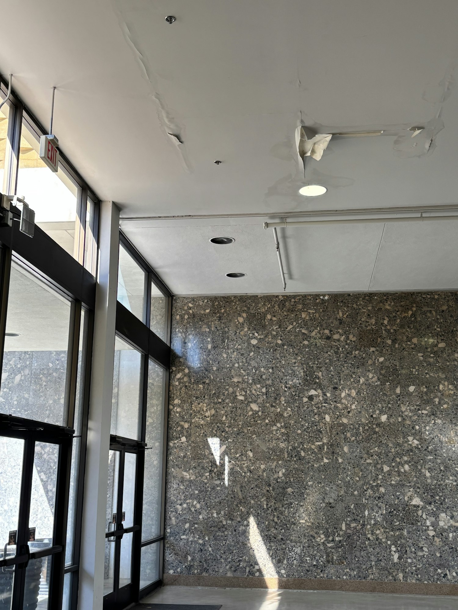 A damaged ceiling with peeling paint, a light fixture, and a terrazzo floor with sunlight streaming through glass doors.