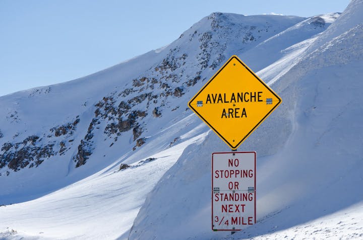 A yellow sign warning of an avalanche area and a red sign prohibiting stopping or standing for the next ¾ mile, set in snowy mountains.