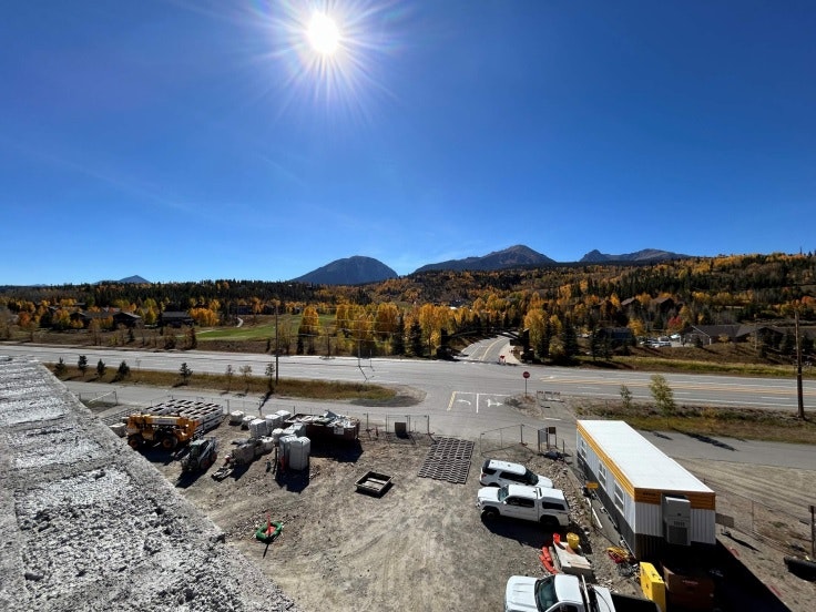 Sunny construction site with vehicles, mountains in the background, and autumn trees.