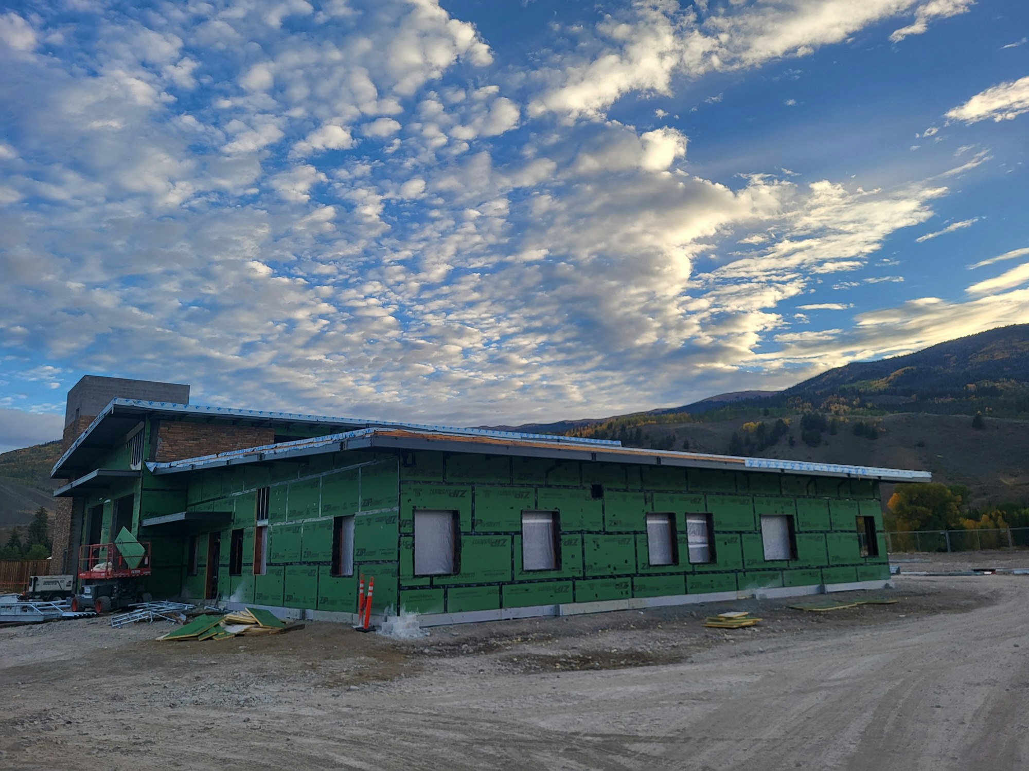 Construction site of a green-wrapped building with mountains and cloudy sky in the background.