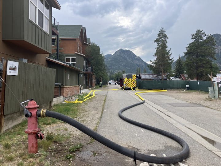 Street view with a fire hydrant connected to a large hose, buildings beside, caution tape ahead, and mountains in the background.