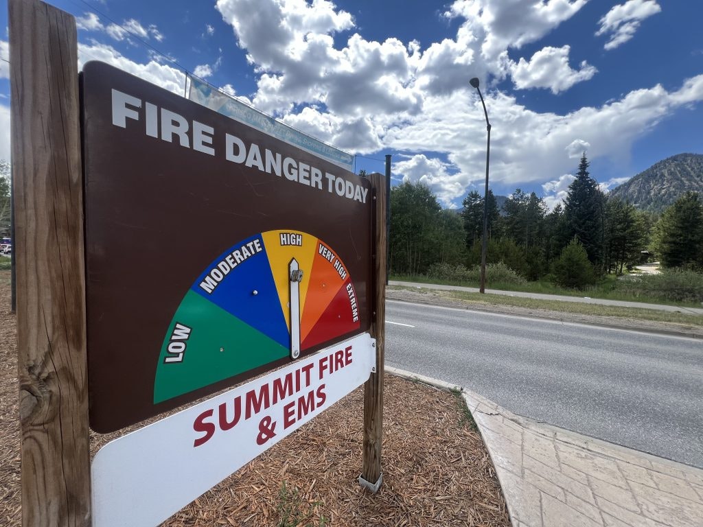 Fire danger sign indicating high risk, by roadside with trees, mountains, and cloudy sky.