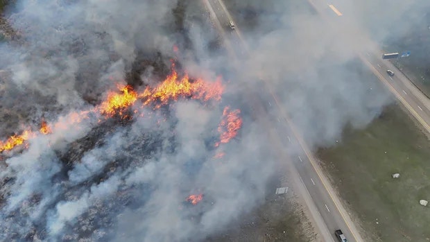 Aerial view of a wildfire next to a highway, with flames and thick smoke visible.