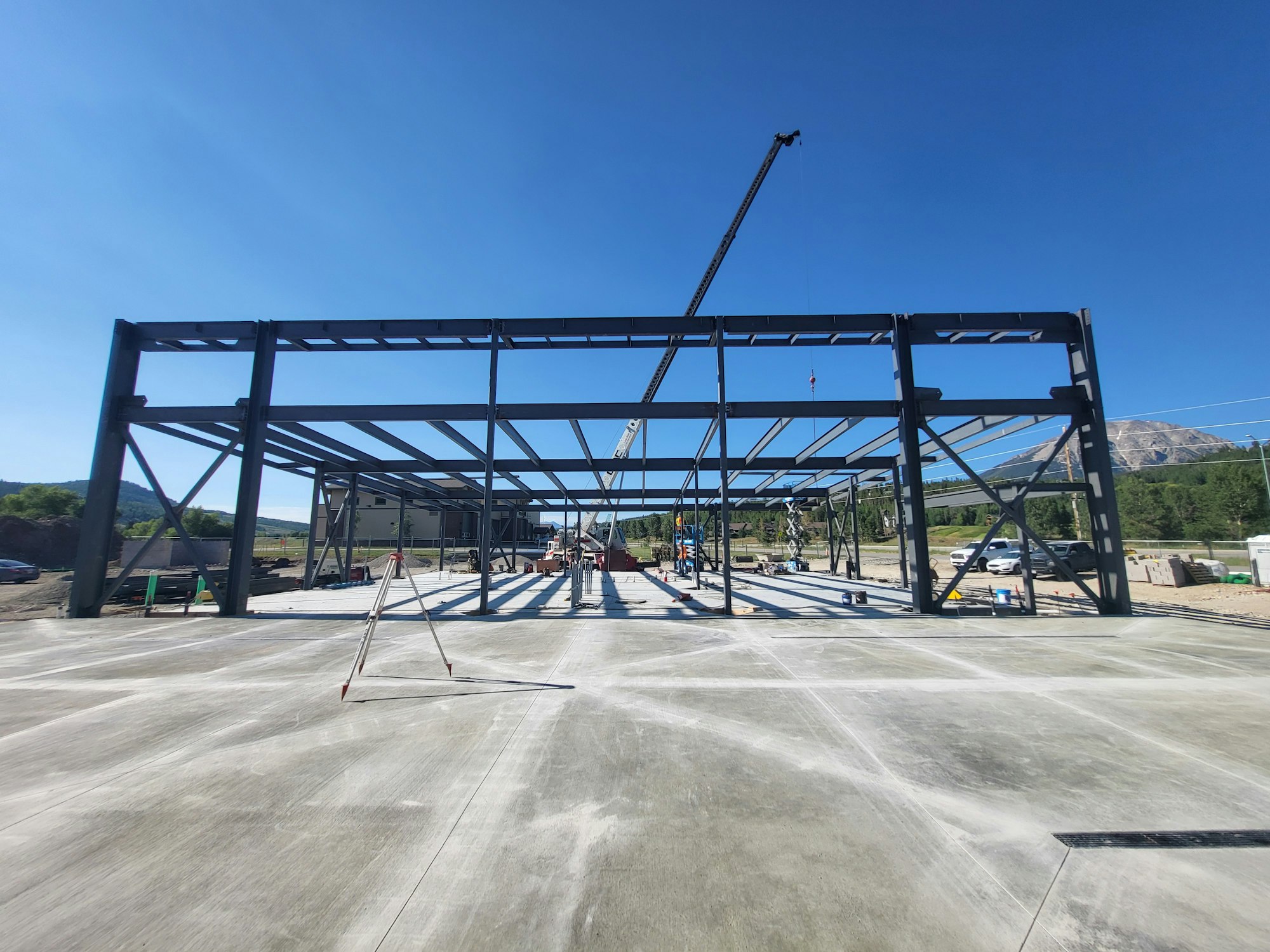 Construction site with steel frame structure, clear sky, crane, workers, and mountains in background.