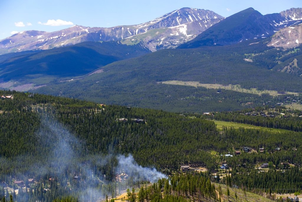 Mountain landscape with forests, scattered homes, and smoke rising near the foreground.