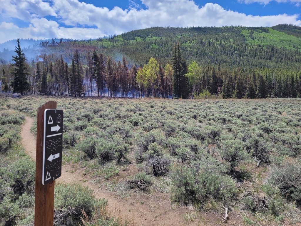 Trailhead sign with directions, footpath, sagebrush, trees, and hills under a partly cloudy sky.