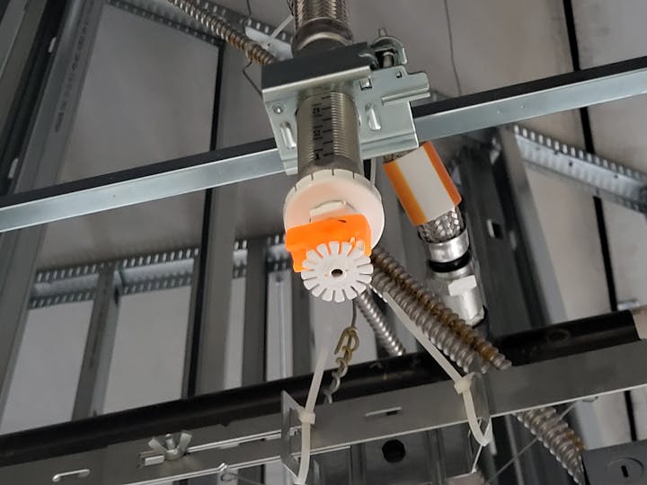 The image shows a fire sprinkler head mounted on a ceiling structure, surrounded by metal supports and piping.