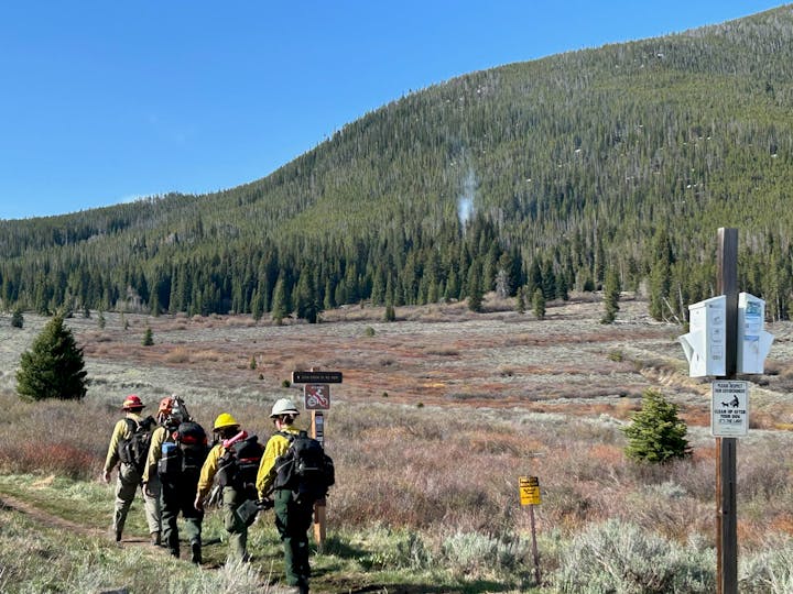 Firefighters in gear walk along a trail in a forested mountain landscape under a clear blue sky.
