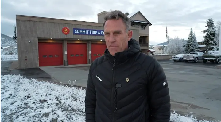 A man stands in front of a fire station with snowy surroundings, wearing a black jacket, looking serious.