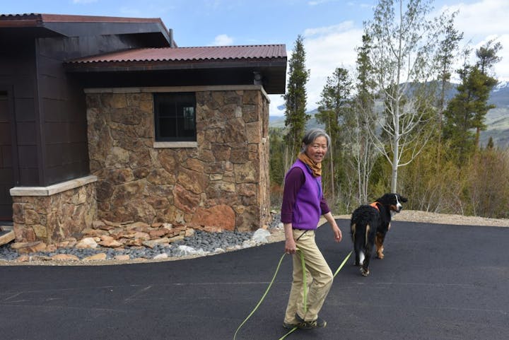 A woman walking a dog on a paved driveway next to a stone house, with trees and mountains in the background.