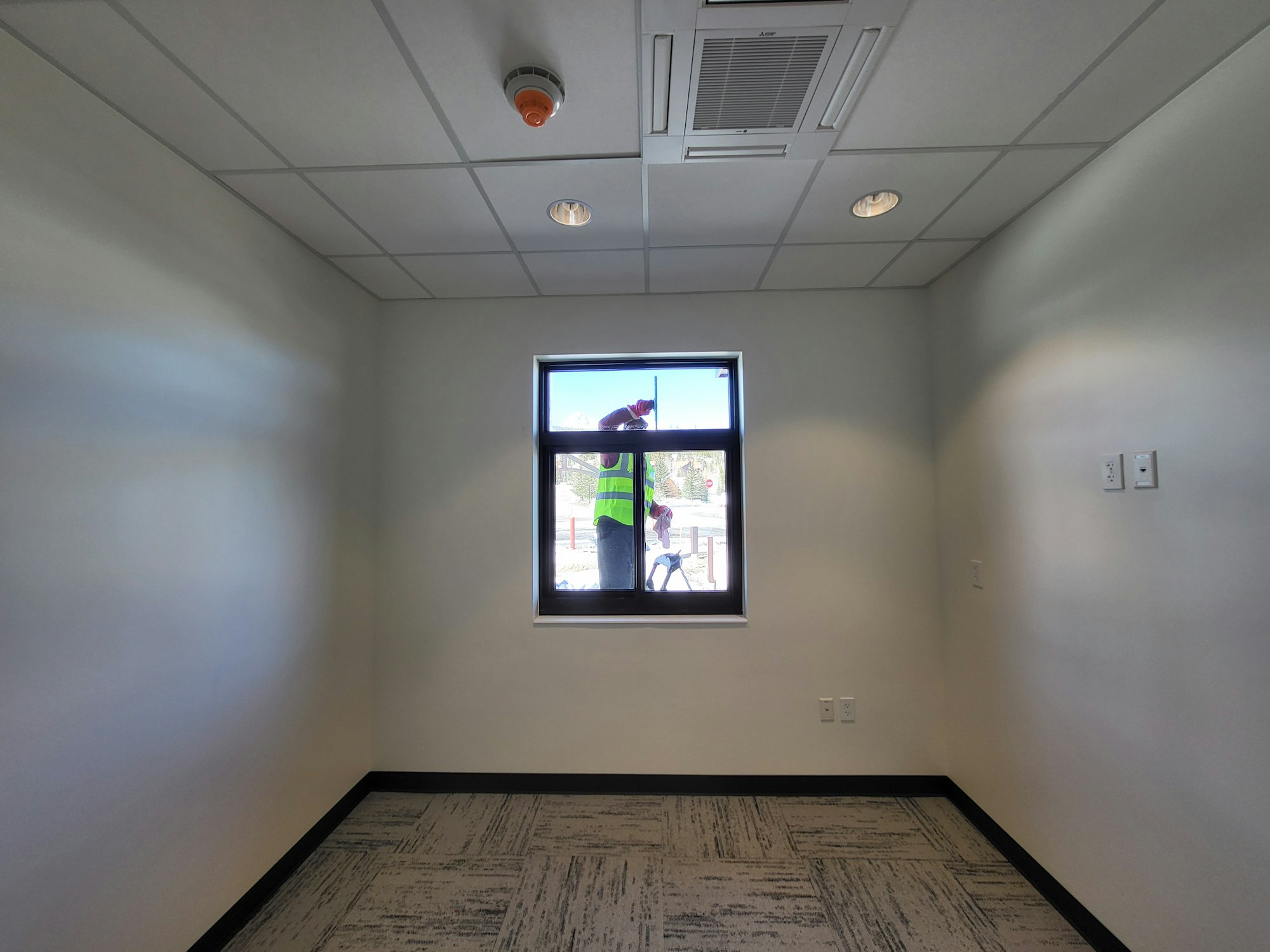 Empty room with gray carpet, white walls, and a ceiling with lights and vents. A person in a safety vest is visible through the window.