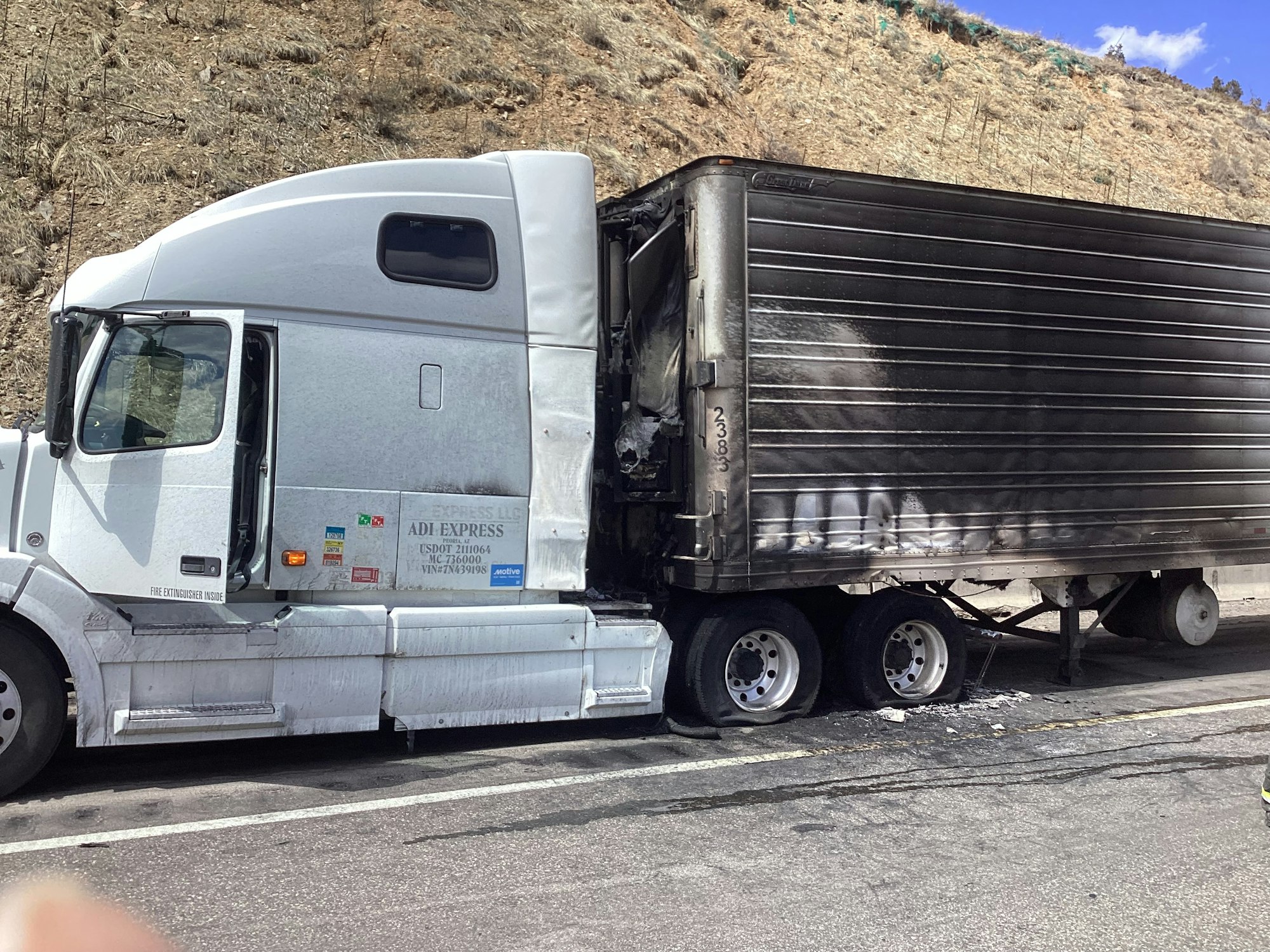 Semi-truck with burnt-out trailer attached, stopped by the roadside.