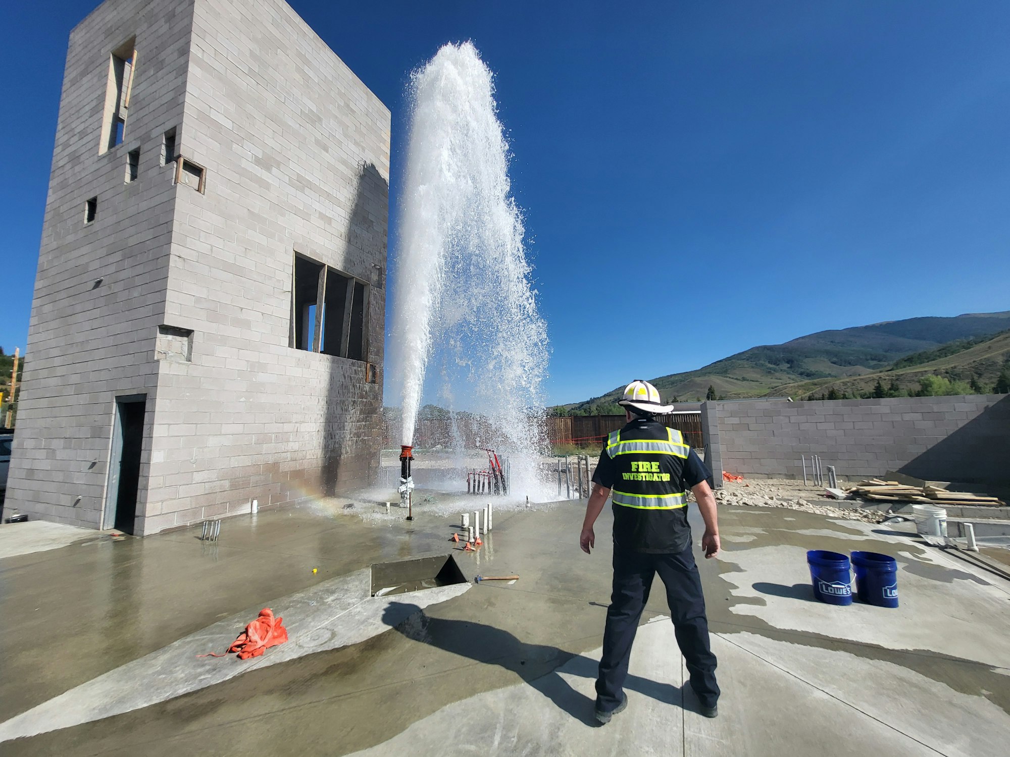 A person in a firefighter's vest labeled "FIRE INVESTIGATOR" watching a high-pressure water leak at a construction site.