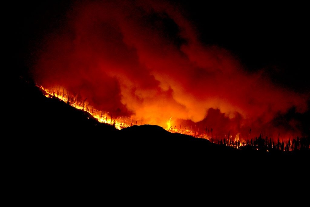 A nighttime wildfire, with vivid flames engulfing a hillside and smoke billowing against a dark sky.
