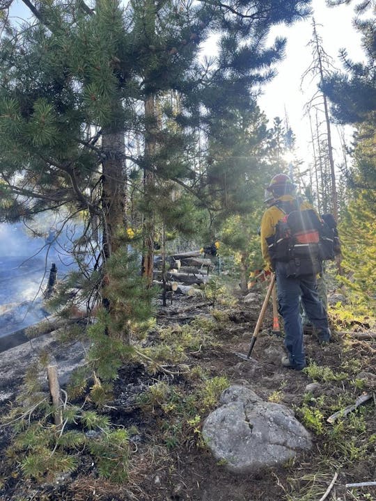 Firefighter in yellow gear with tools in a forested area with signs of fire.