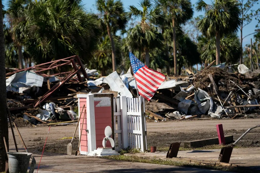 An American flag stands amid destruction, possibly from a disaster, with debris and toppled structures surrounded by palm trees.