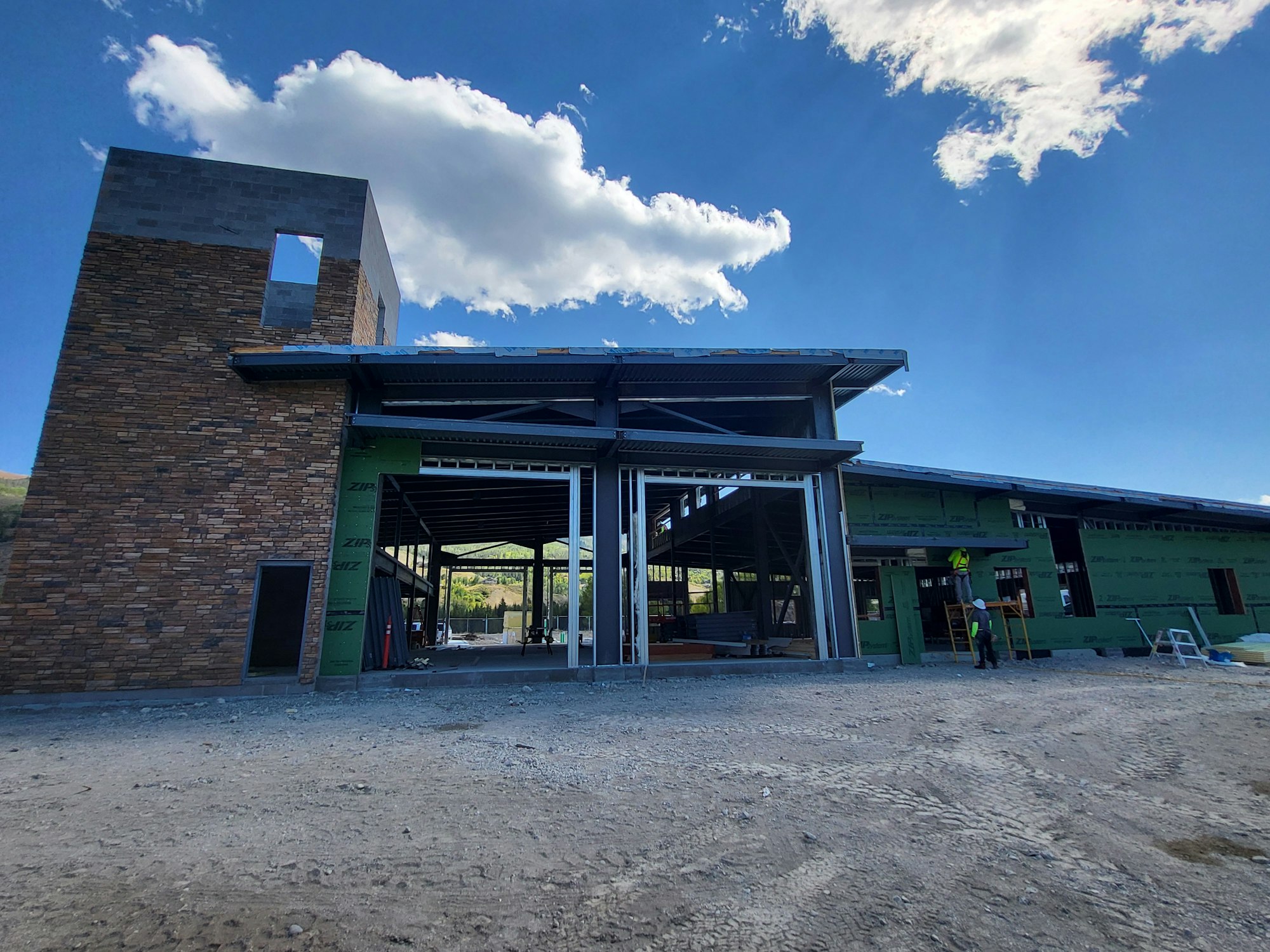 Construction site with unfinished building, worker in high-vis gear, and clear blue sky with clouds.