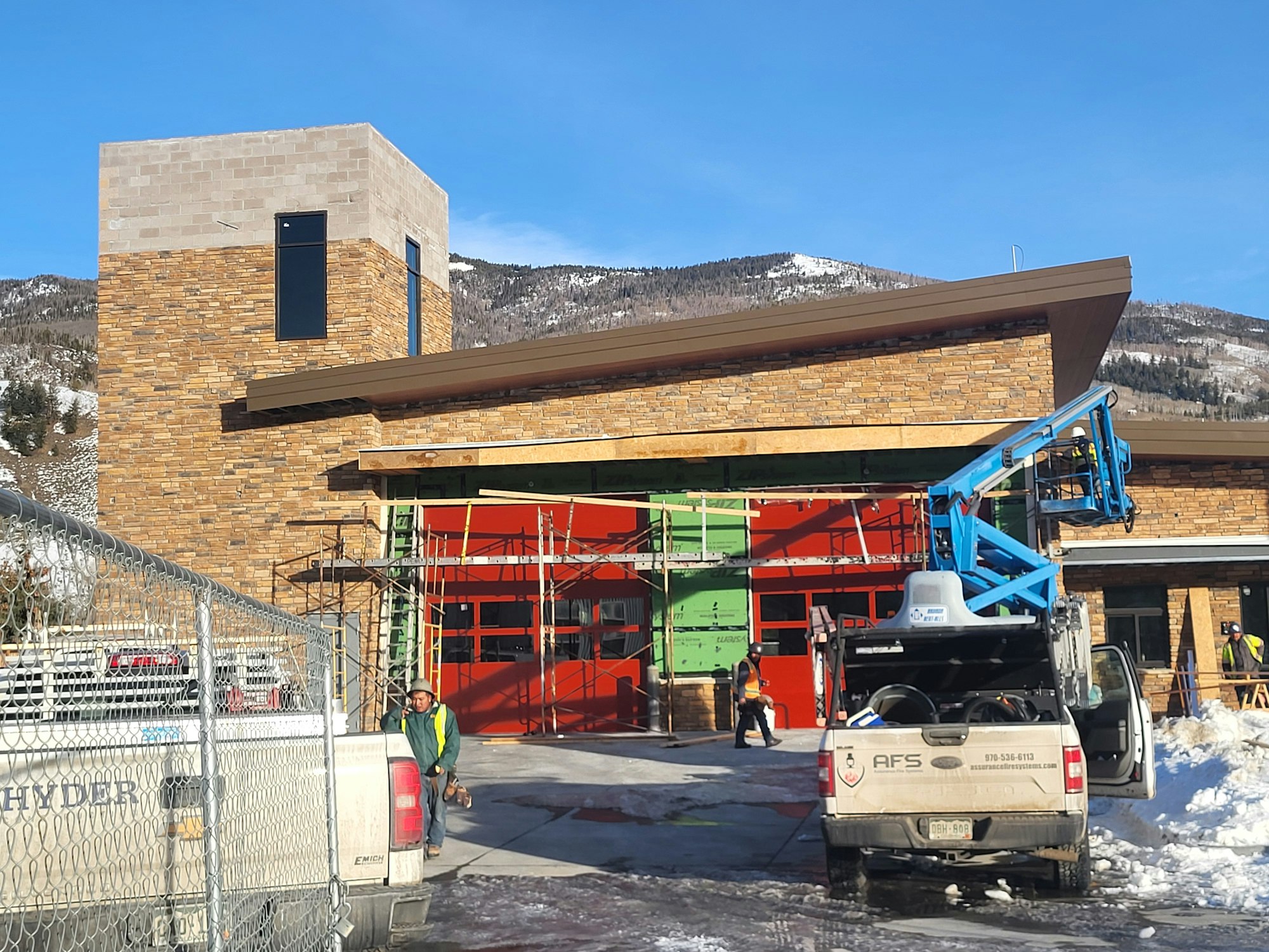 A construction site with brick building, vehicles, a blue lift, and workers in a snowy landscape.