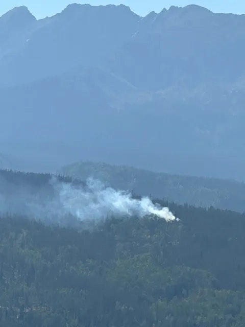 Wildfire smoke rising from a forest with mountain backdrop.