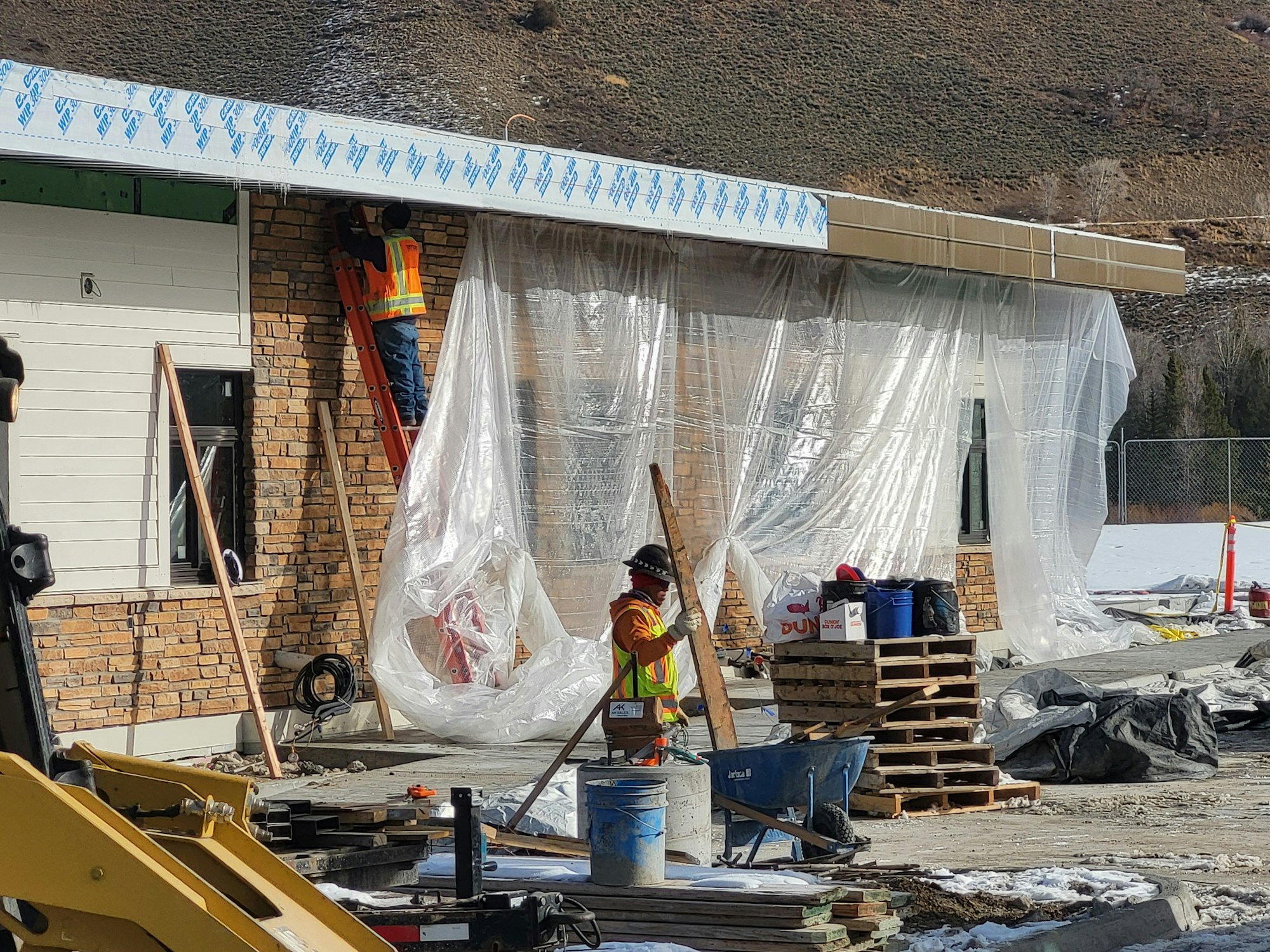 Construction site with a worker on a ladder, plastic sheeting, brick wall, tools, and building materials.