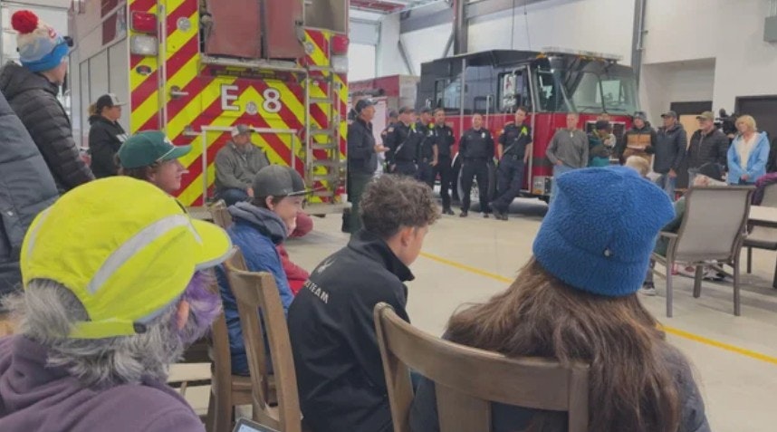 A group of people is gathered inside a fire station, with firefighters present and a fire truck in the background.