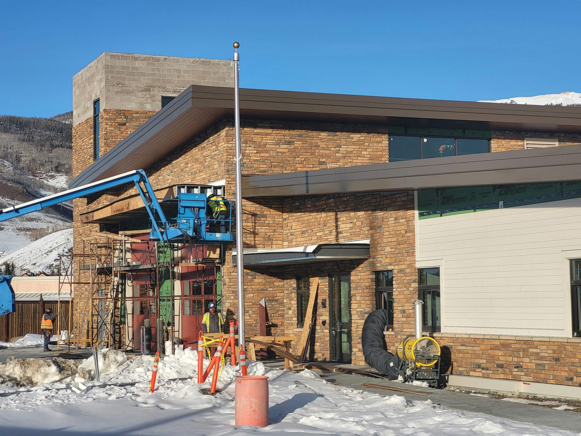A building under construction with workers, scaffolding, a lift, and snow on the ground.