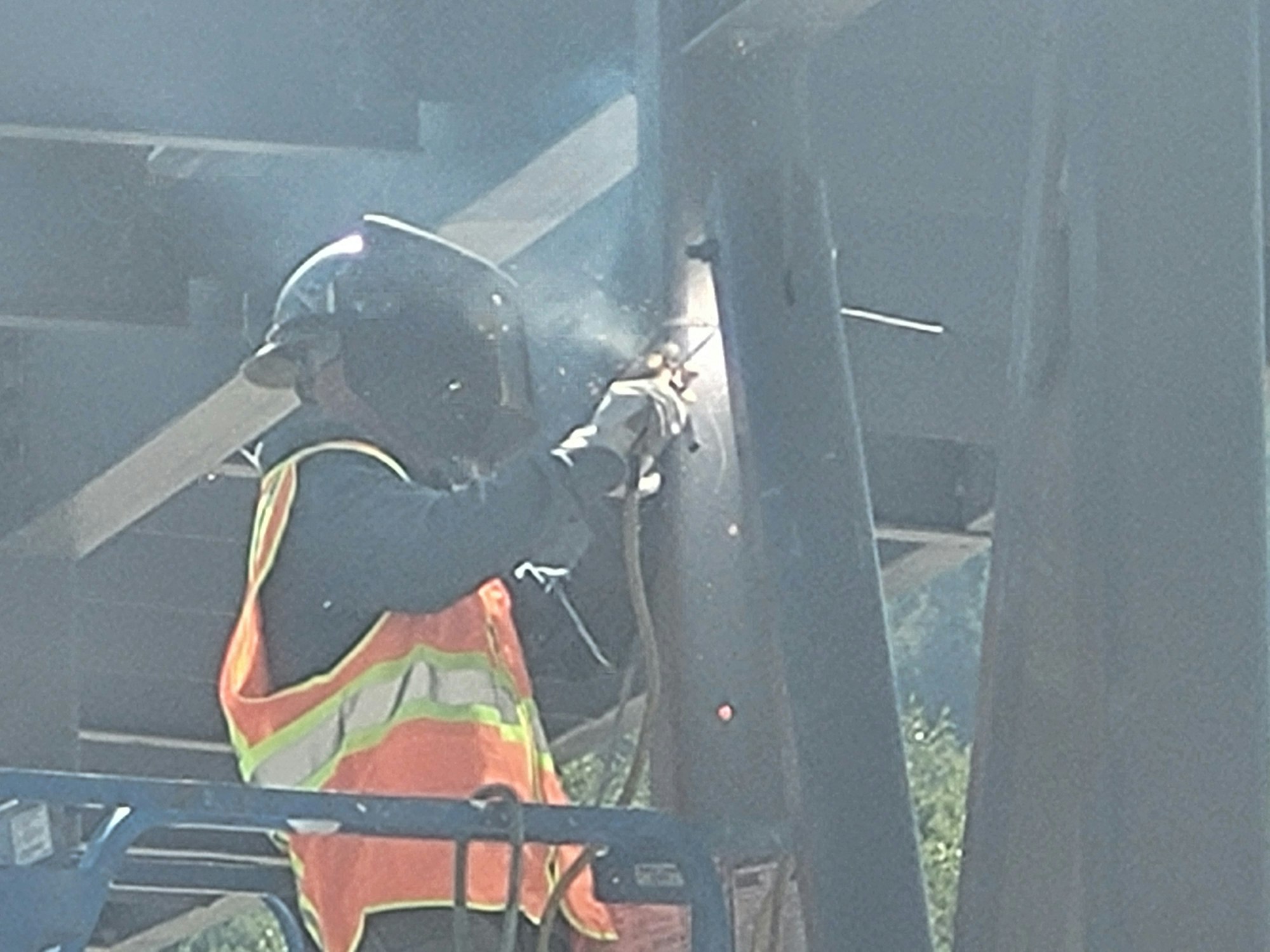 A worker in high-visibility gear is welding metal outdoors, with visible sparks.