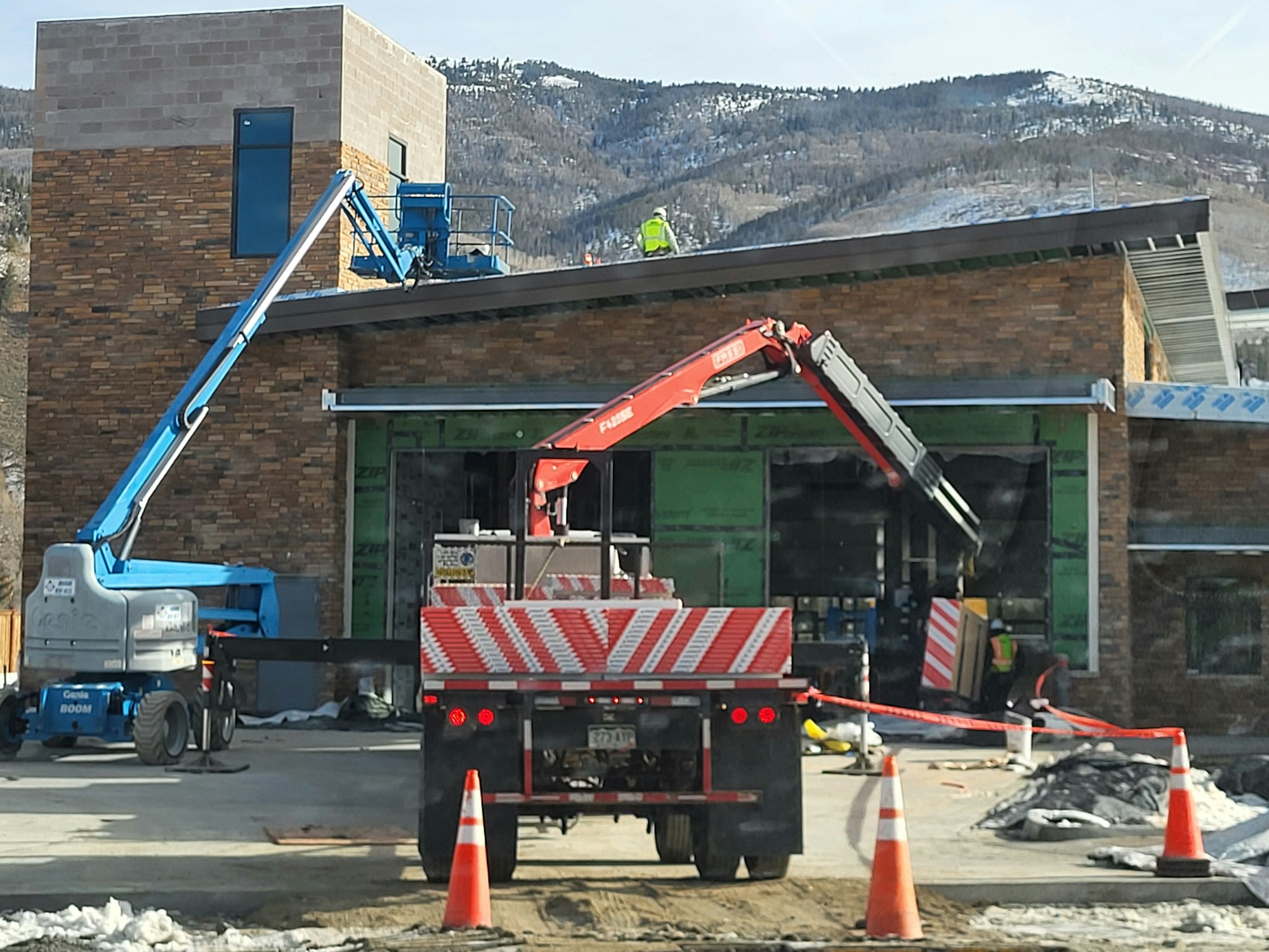 Construction site with machinery, including a boom lift and excavator, with mountains in the background.
