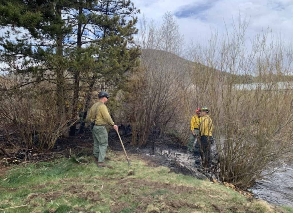 Three people in safety gear manage a small, burnt area in a grassy, wooded environment.