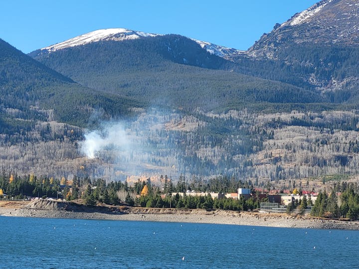 A mountainous landscape with a lake in the foreground, smoke rising from trees, and snow-capped peaks in the background.