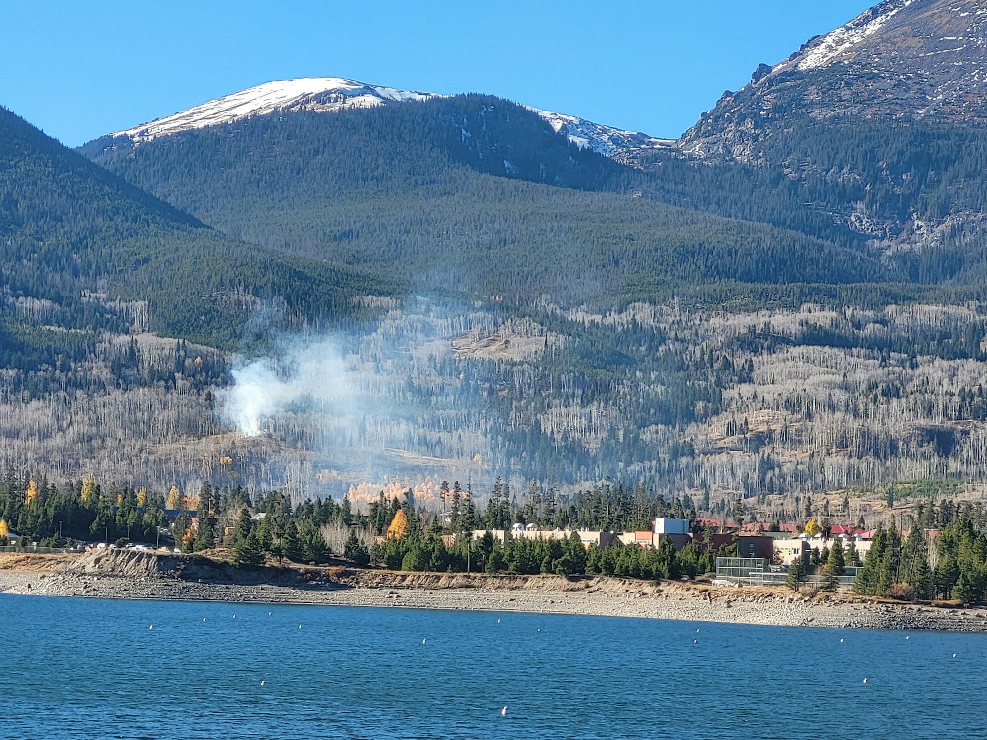 A mountainous landscape with a lake in the foreground, smoke rising from trees, and snow-capped peaks in the background.