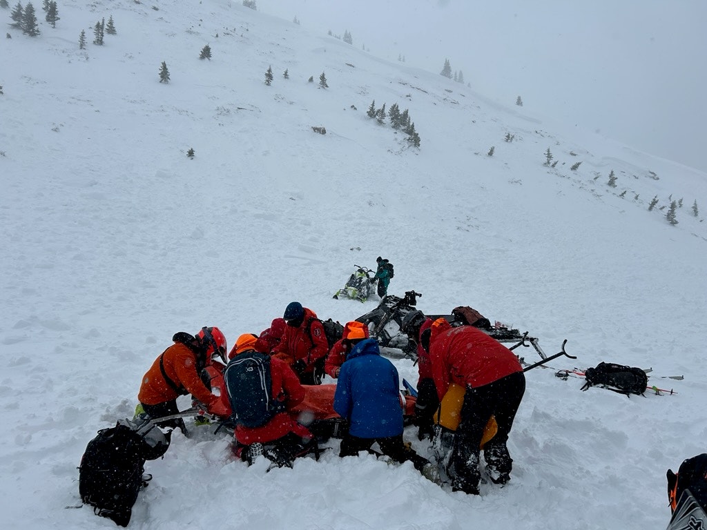 A group of people in winter gear gather around snowmobiles on a snowy slope.