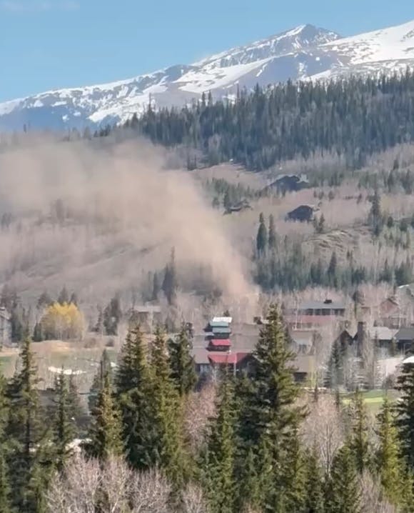 A structure in a forested area emits smoke or dust, with mountains in the background.