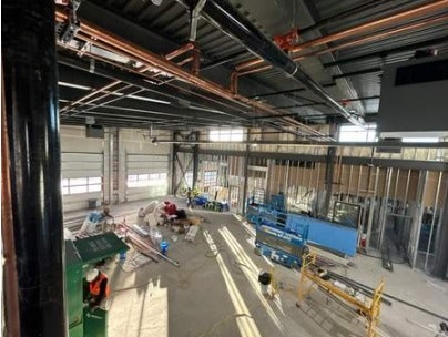 Construction site interior with exposed pipes above, materials on floor, and workers in high-visibility vests.