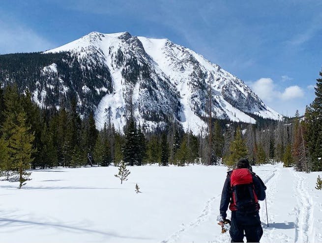 A hiker walks in snow towards a mountain under a clear blue sky.