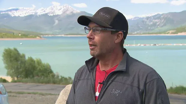 A man wearing a hat and glasses stands by a lake, with mountains in the background and clear skies above.