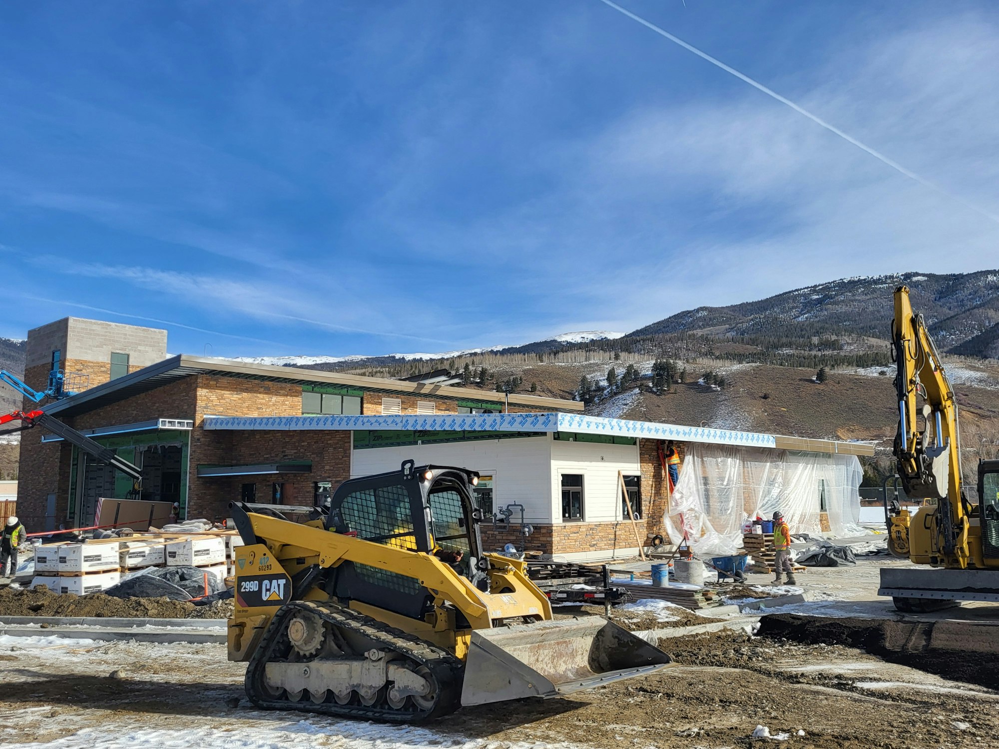 Construction site with machinery, building, workers, and snowy mountains in the background.