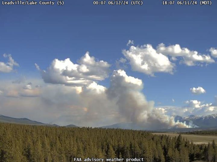 A forested landscape with a large smoke plume, possibly from a wildfire, under a cloudy sky.