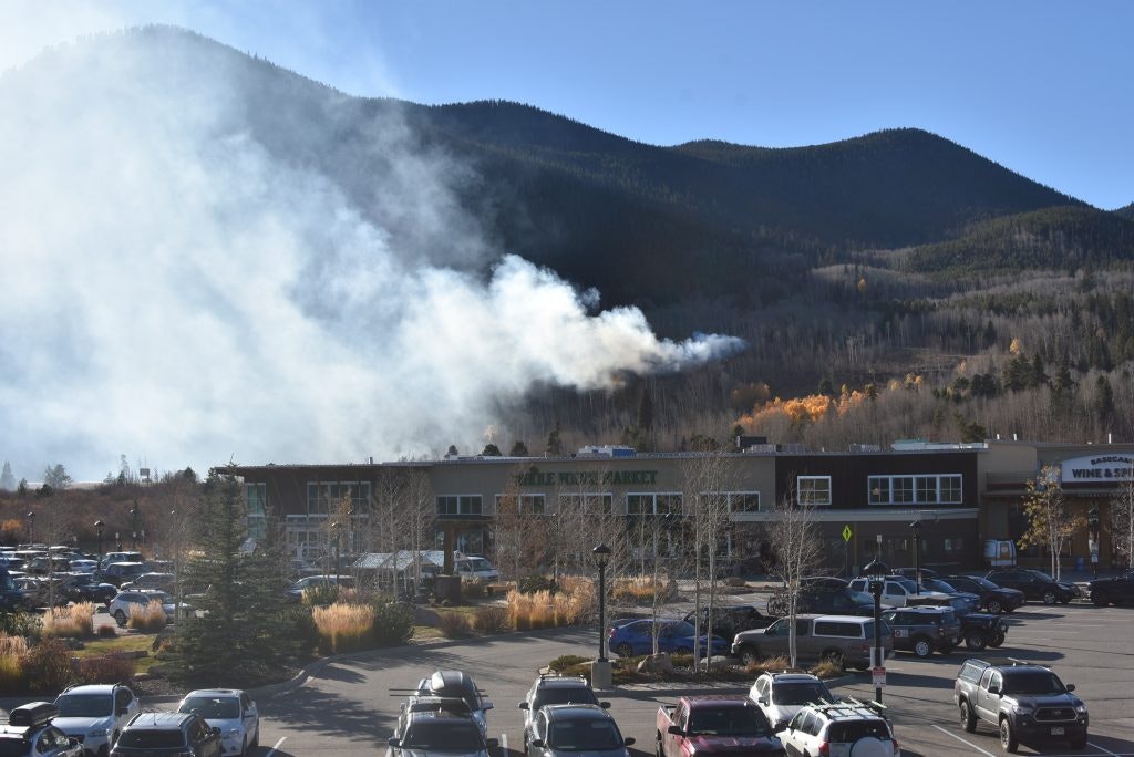 A parking lot with cars in front of stores, smoke rising from trees on a mountain in the background.