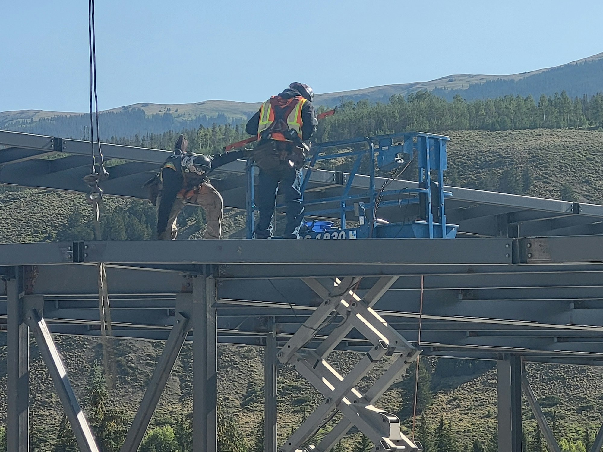 Two workers on a lift working on a steel structure with nature in the background.