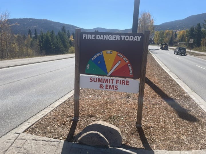 Fire danger sign next to a road, indicating "very high" risk, with forested hills and vehicles in the background.